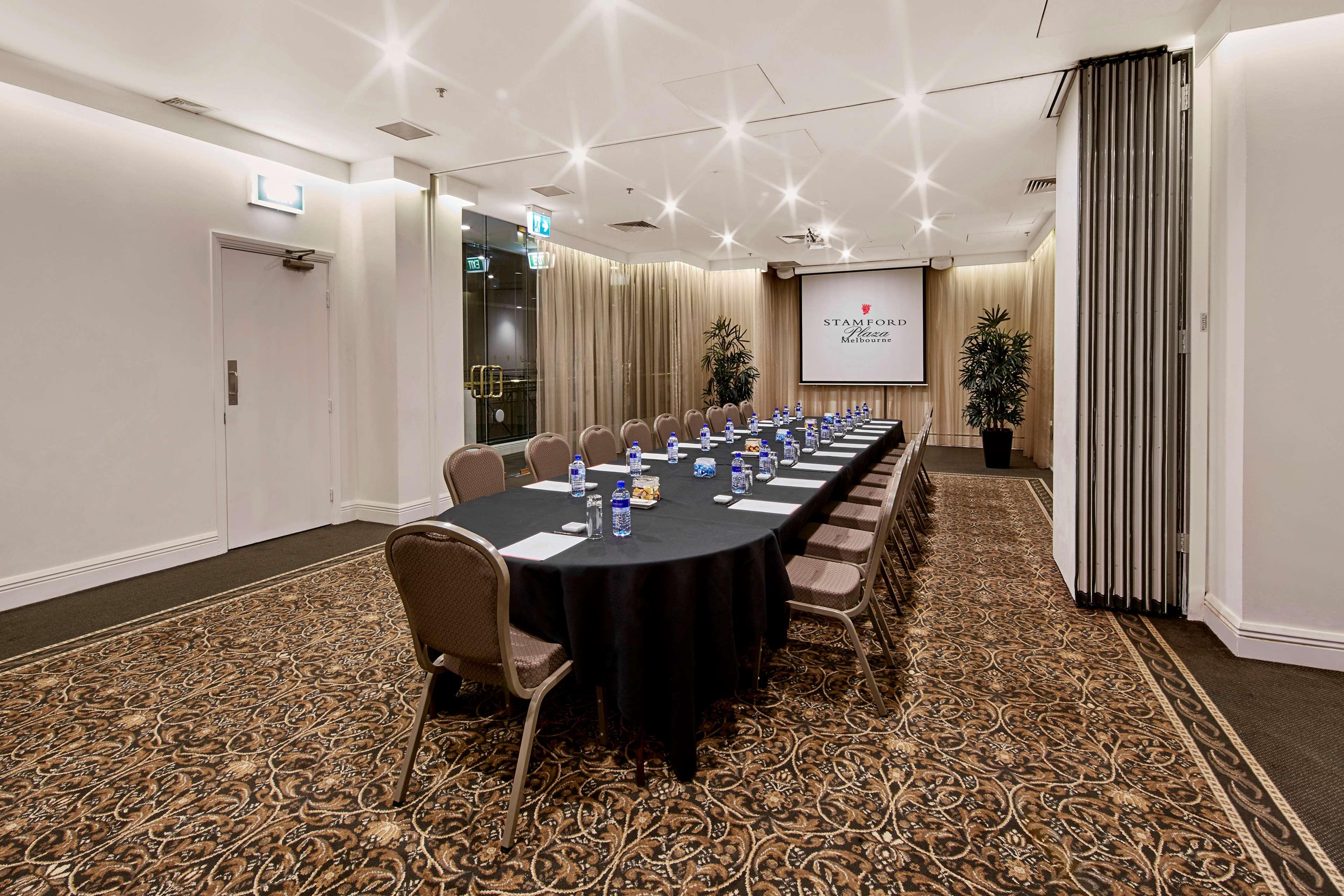 Hotel Stamford Plaza Melburne Exterior foto The photo shows a meeting room set up for a conference or presentation. There is a long, rectangular table covered with a black tablecloth, surrounded by chairs. Each seat has a water bottle placed in front of it. At the front of the room, there is a