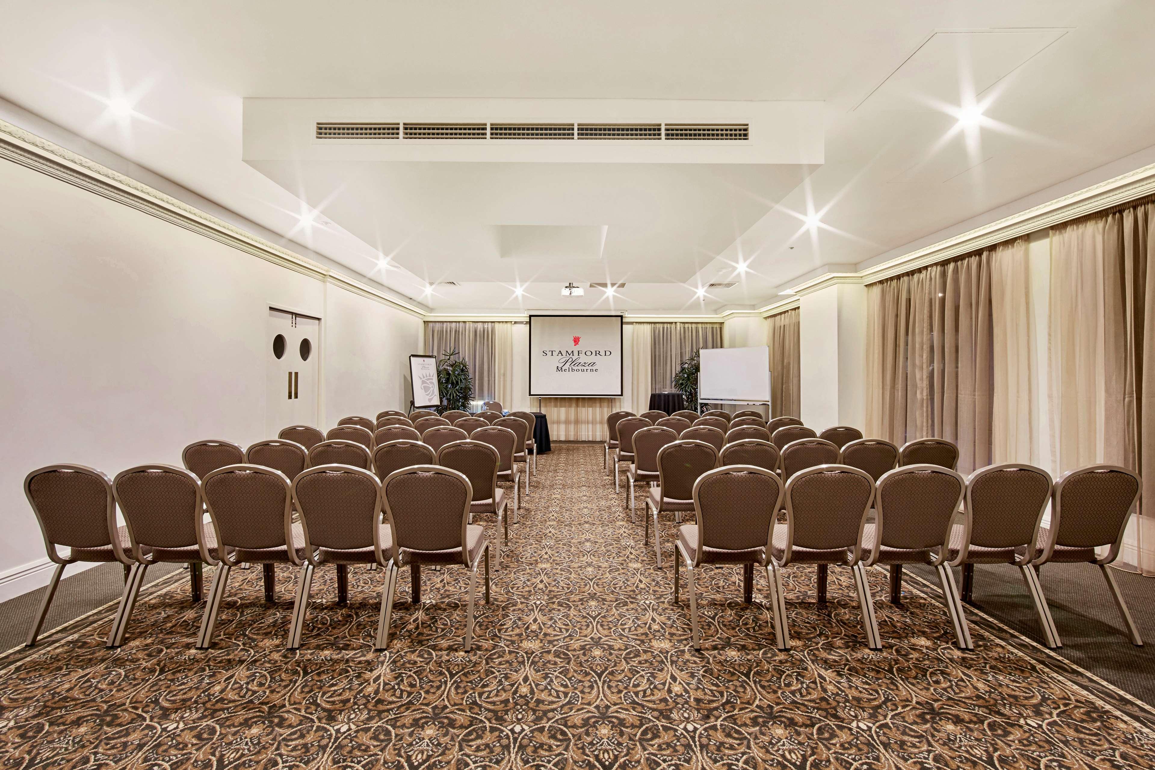 Hotel Stamford Plaza Melburne Exterior foto The photo shows a conference room set up for an event. It features several rows of beige chairs arranged facing a central presentation area. At the front, there is a screen displaying a logo or title, possibly for a conference or meeting. The walls h