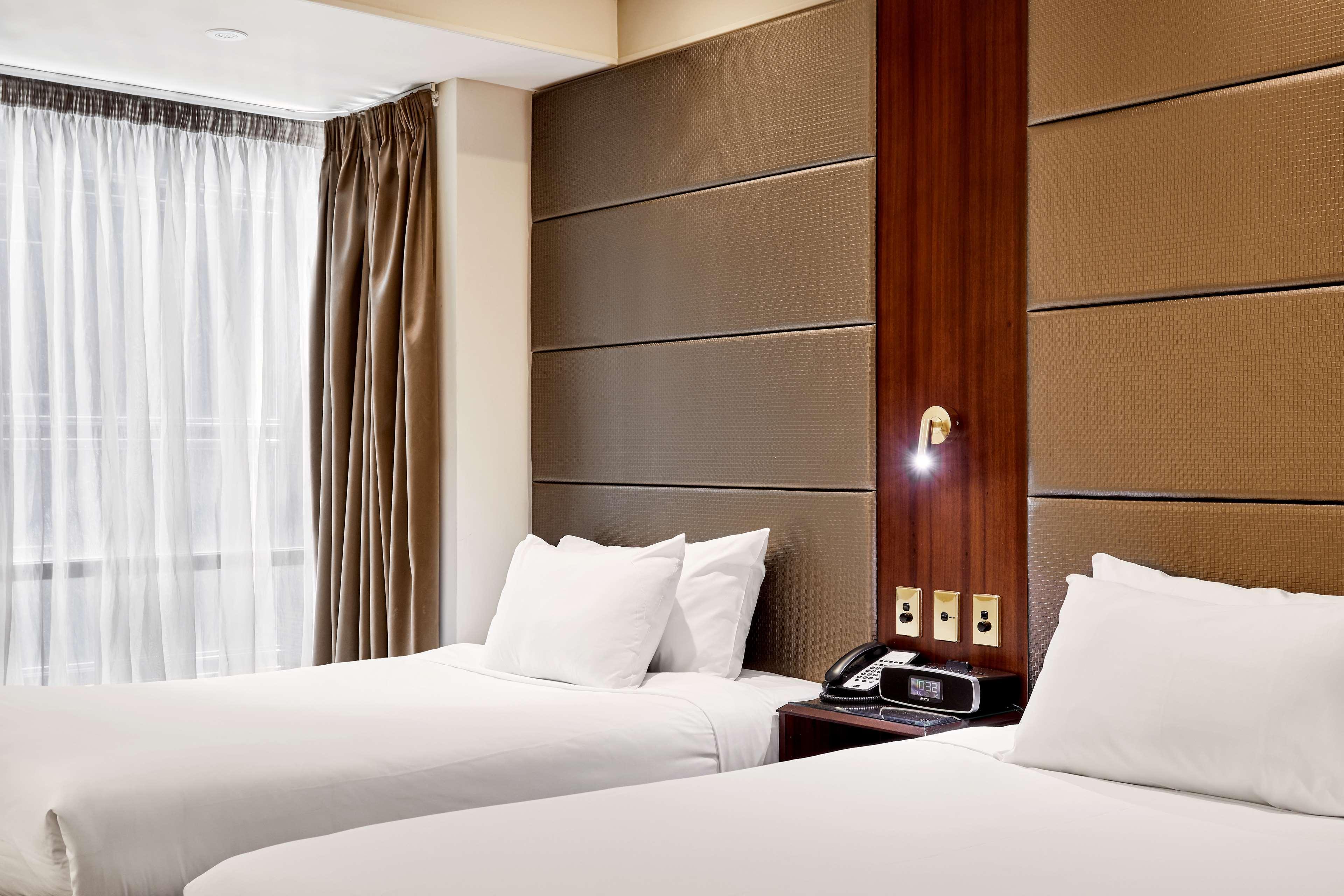 Hotel Stamford Plaza Melburne Exterior foto The photo shows a modern hotel room featuring two twin beds with white linens and fluffy pillows. The walls behind the beds have a combination of textured panels in neutral colors. There is a bedside table with a lamp and a phone. A large window with