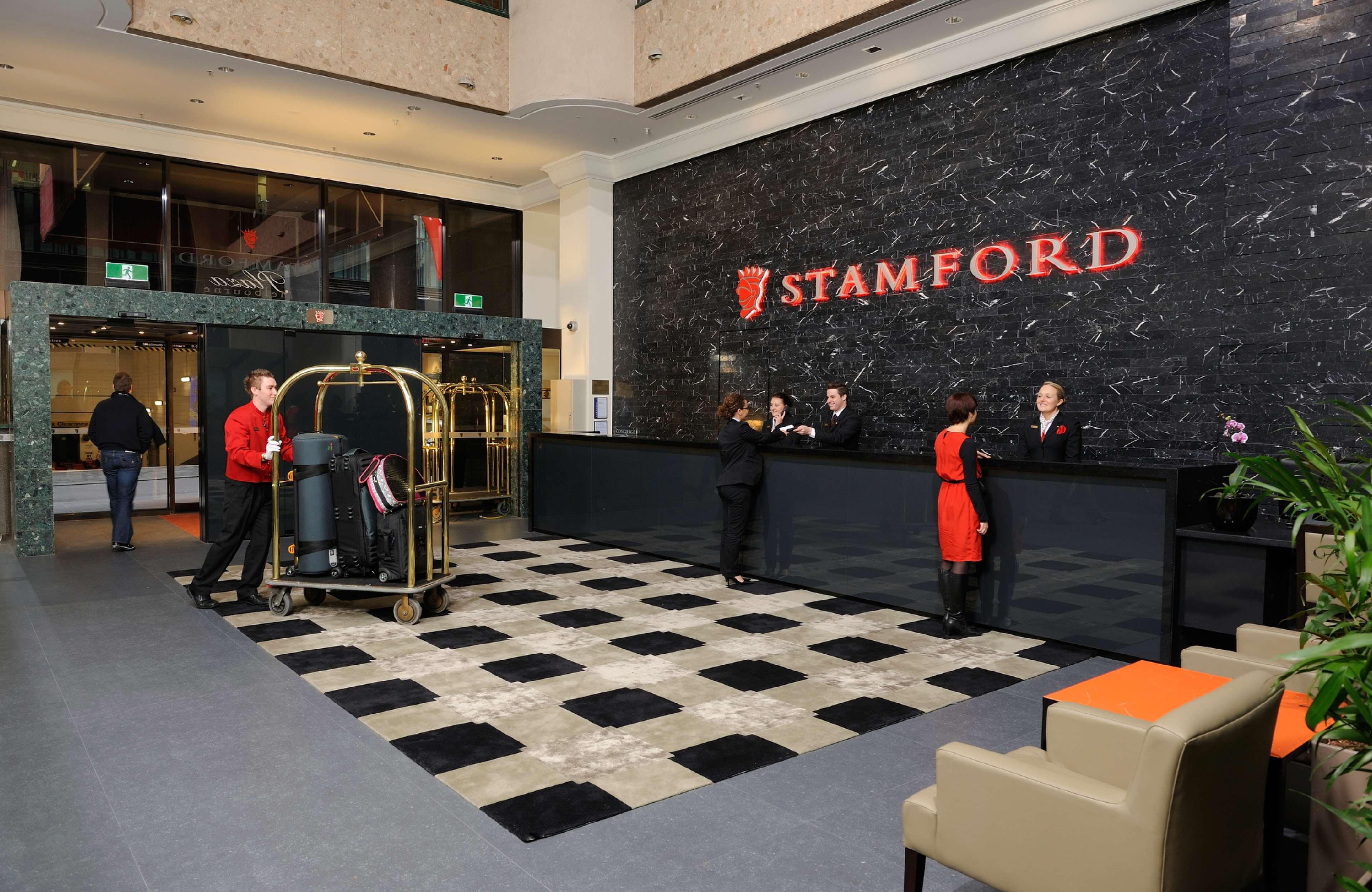 Hotel Stamford Plaza Melburne Interior foto The photo shows the interior of a hotel lobby, specifically the Stamford Hotel. There is a large reception desk where several staff members are attending to guests. In the foreground, a bell cart loaded with luggage is positioned beside the reception