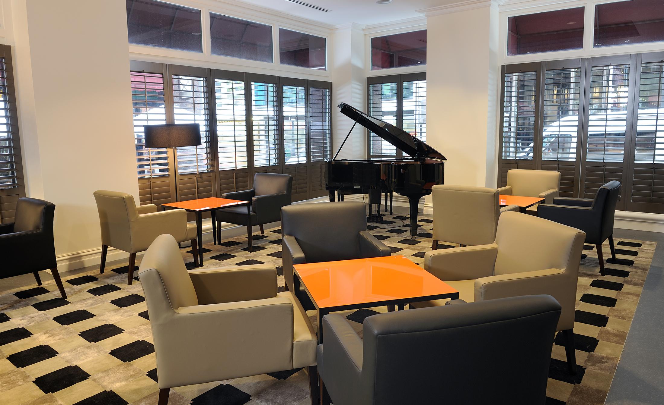 Hotel Stamford Plaza Melburne Exterior foto The photo shows a stylish indoor lounge area. It features several light gray armchairs arranged around orange square tables. In the corner, there is a black grand piano, adding an elegant touch to the space. The room has large windows with wooden shu