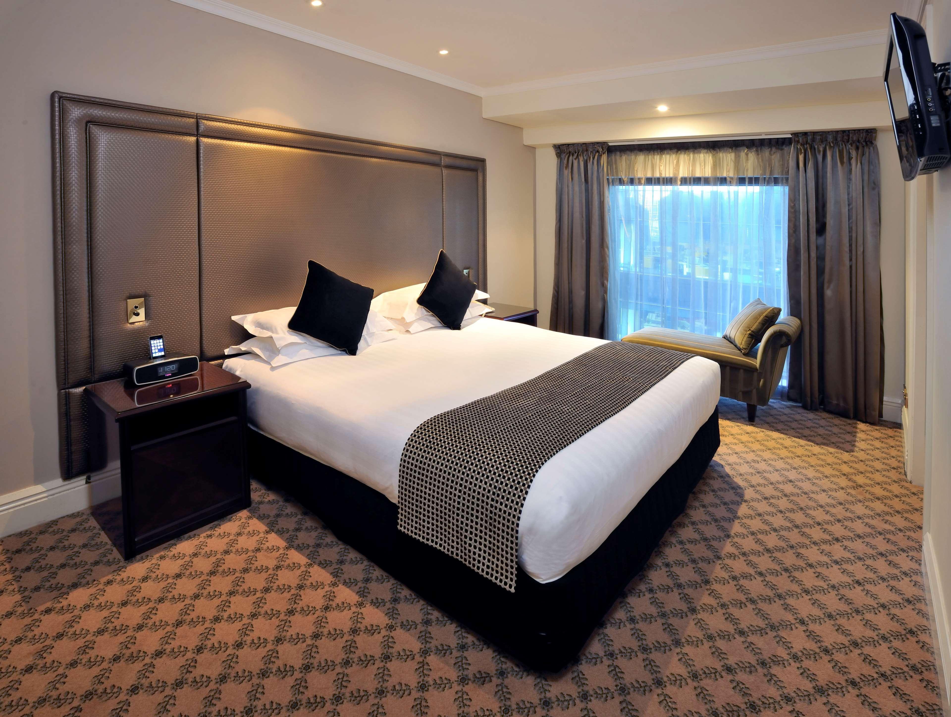 Hotel Stamford Plaza Melburne Exterior foto The photo shows a hotel room featuring a neatly made king-sized bed with dark pillows. The headboard is upholstered in a dark material, and there is a decorative blanket at the foot of the bed. Beside the bed are two nightstands, each equipped with l