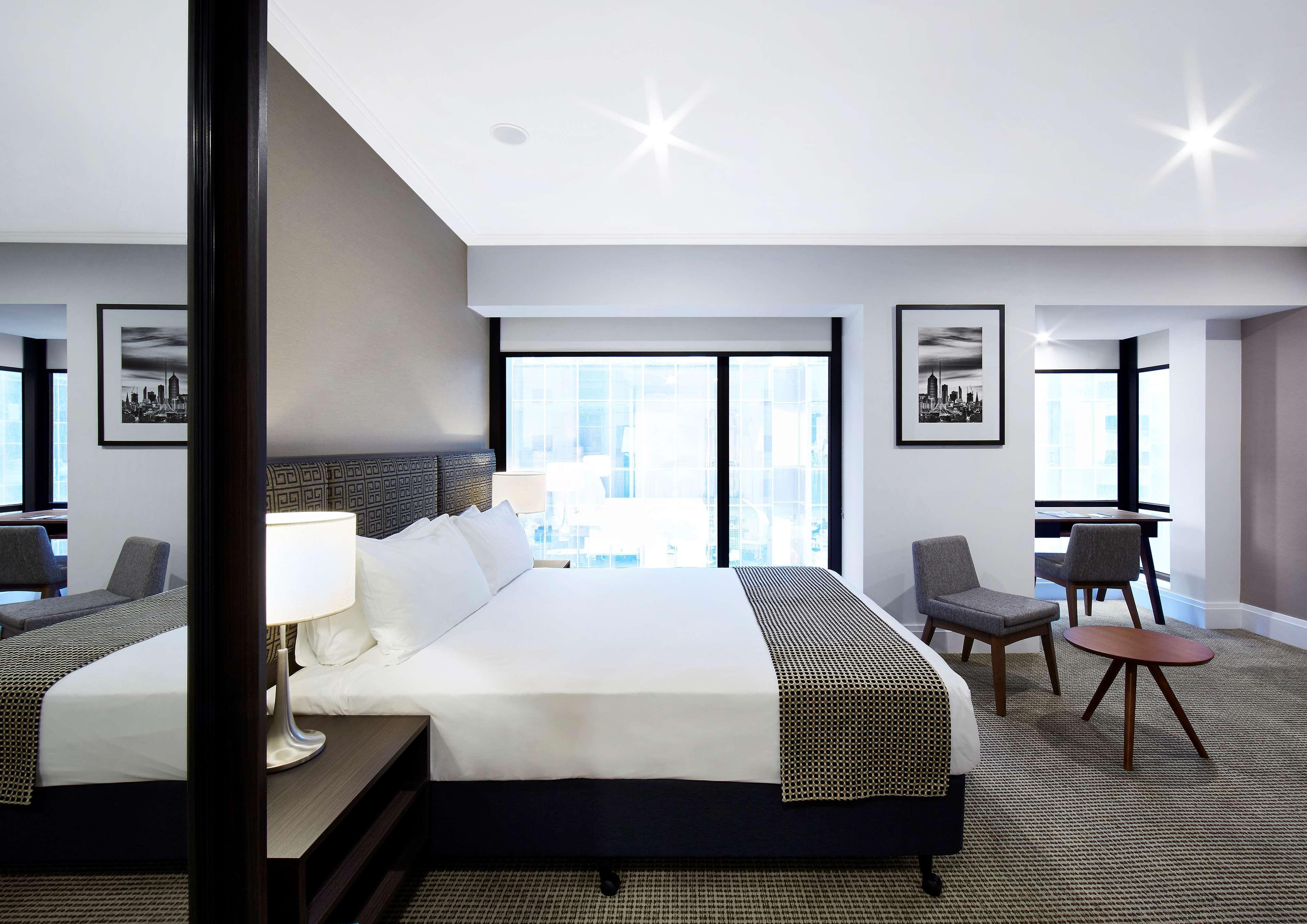 Hotel Stamford Plaza Melburne Exterior foto The image shows a modern hotel room featuring a large bed with white linens and a gray throw blanket. There are two windows that allow natural light to enter the space, and the walls are painted in a light color. In addition to the bed, there are two