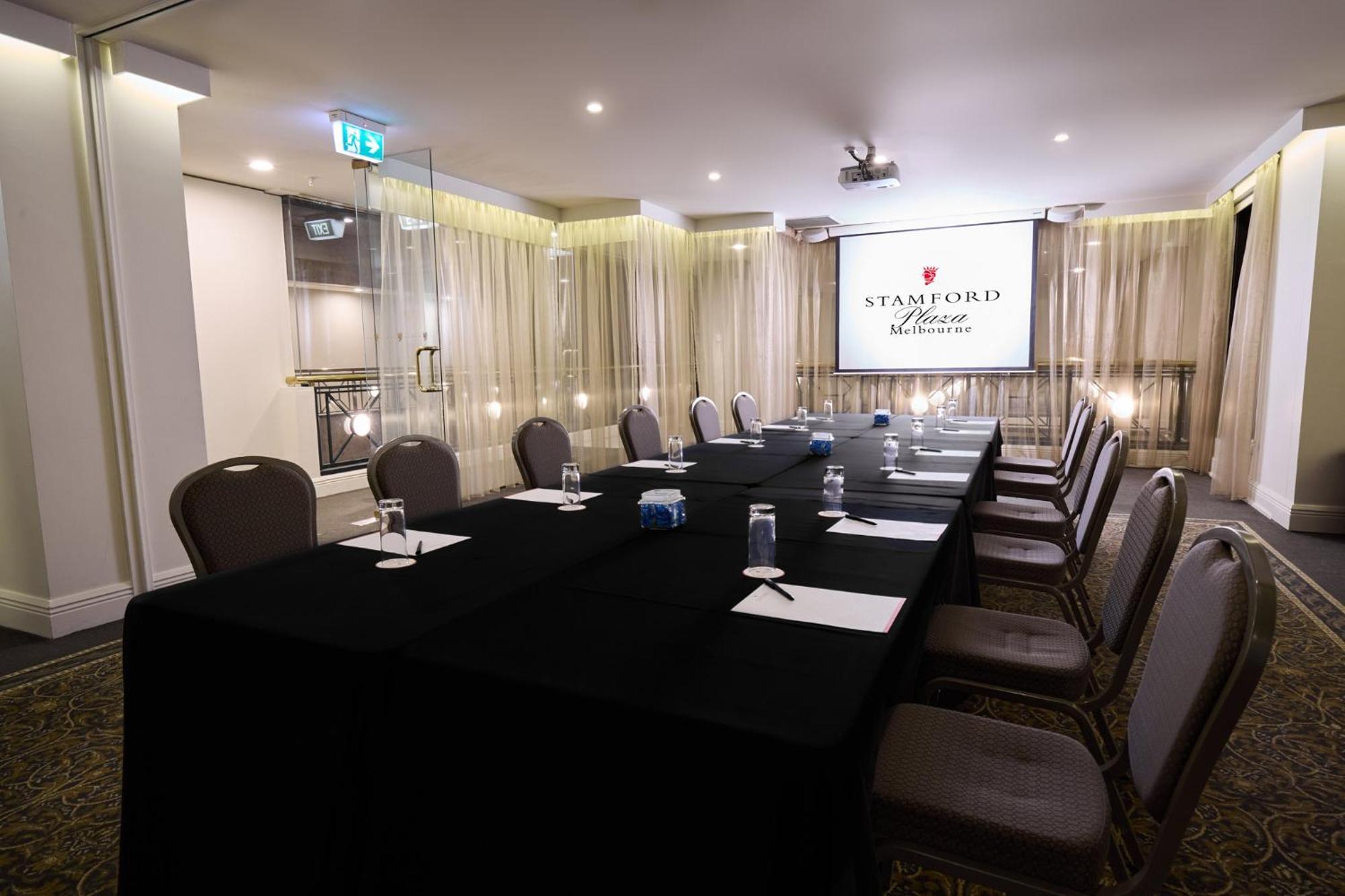 Hotel Stamford Plaza Melburne Exterior foto The photo shows a well-appointed conference room designed for meetings or presentations. There is a long rectangular table covered with a black tablecloth, surrounded by upholstered chairs. On the table, there are several folded pieces of paper and g