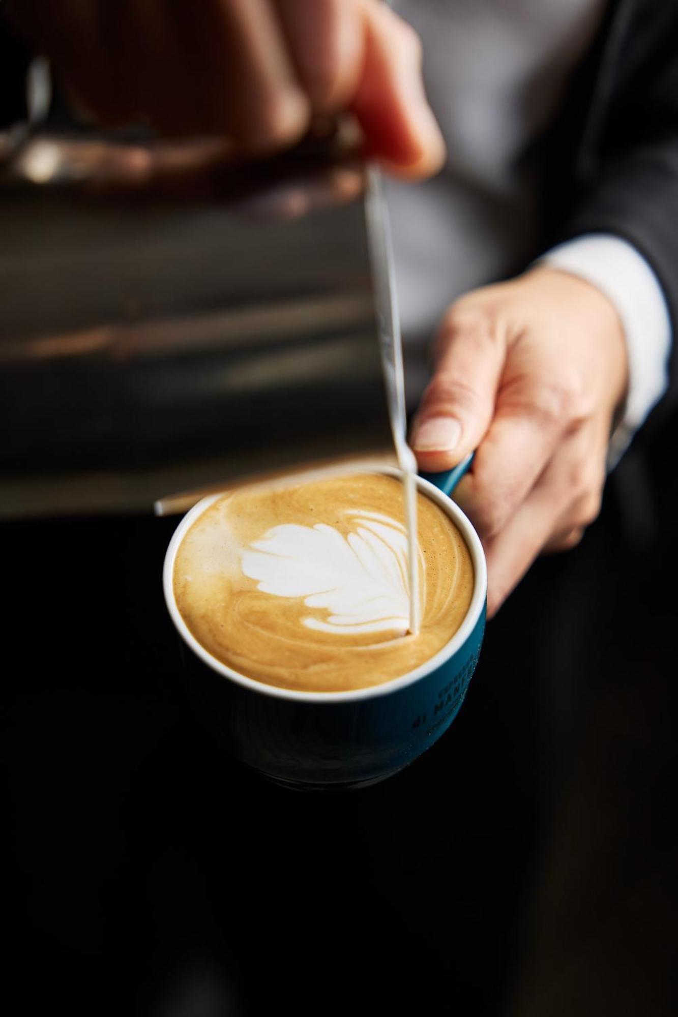 Hotel Stamford Plaza Melburne Exterior foto The photo shows a close-up of a hand holding a cup of coffee with a latte art design on the surface. The coffee appears to be a creamy, caramel color, likely a latte or cappuccino. A small stream of milk is being poured into the cup, creating a heart