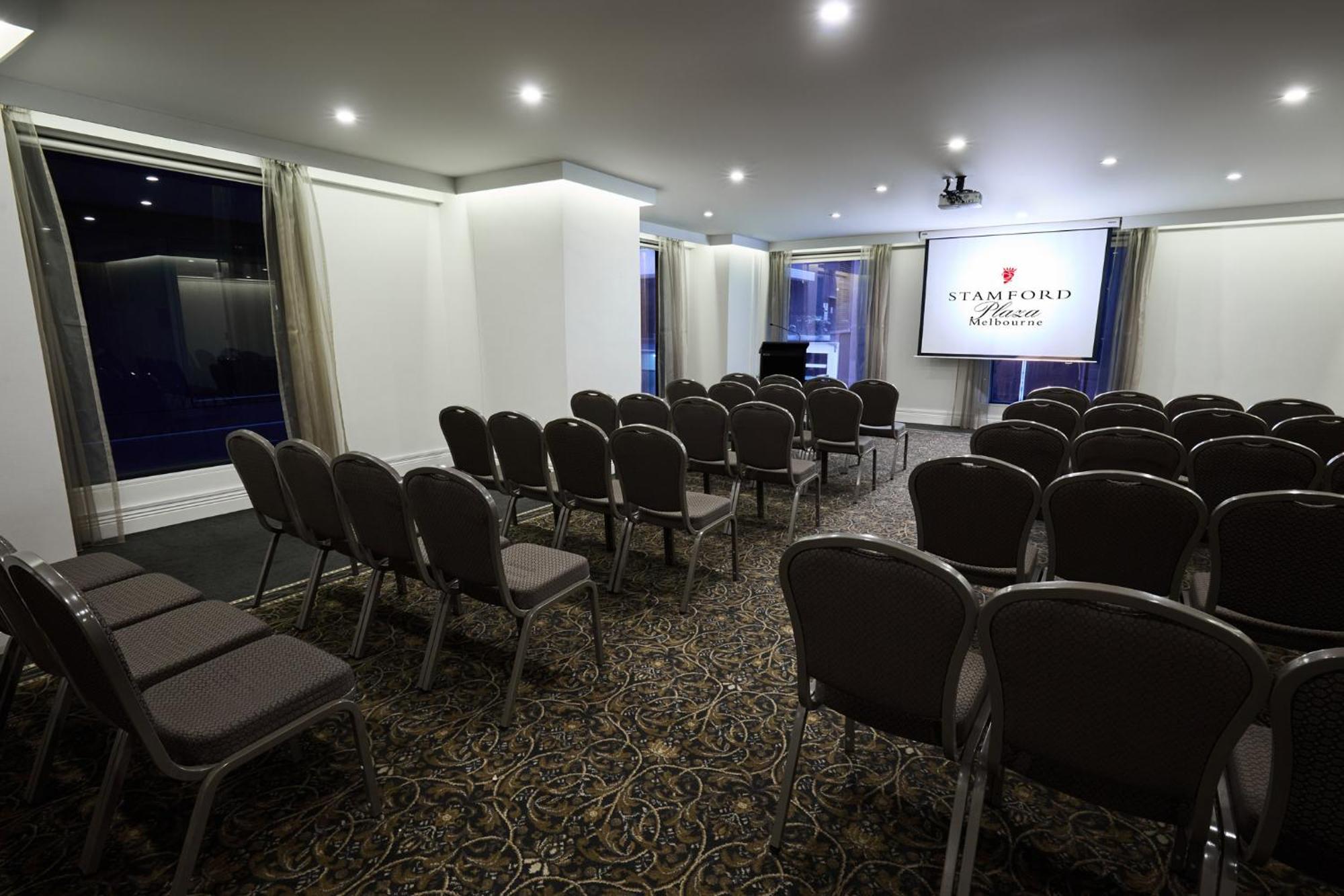 Hotel Stamford Plaza Melburne Exterior foto The image shows a conference or meeting room set up for an event. There are rows of gray chairs arranged facing a large screen displaying the name "Warford House." The room is well-lit with overhead lighting and has large windows covered with light-c