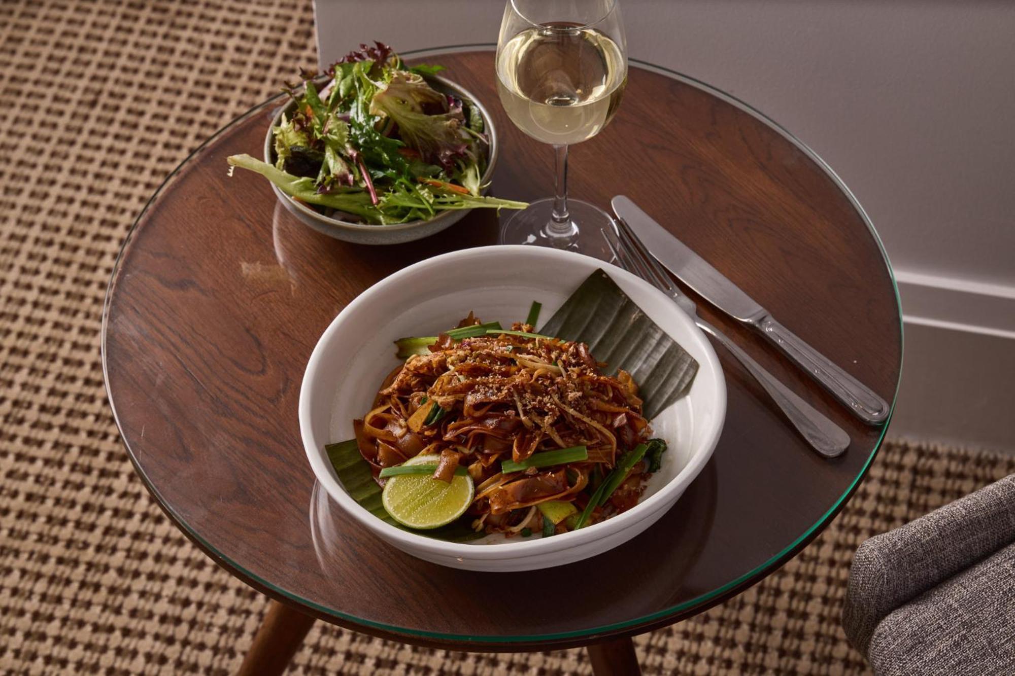 Hotel Stamford Plaza Melburne Exterior foto The photo shows a meal served on a wooden table. In the foreground, there is a bowl of stir-fried noodles garnished with green onions and a lime wedge, set atop a banana leaf. Next to it, there is a bowl of mixed green salad. A glass of white wine si