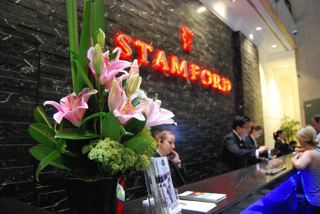 Hotel Stamford Plaza Melburne Exterior foto The photo shows a hotel reception area with a prominent sign that reads "STAMFORD" in bright red letters. In the foreground, there is a decorative arrangement of flowers, primarily featuring pink lilies, along with greenery. The reception counter is 